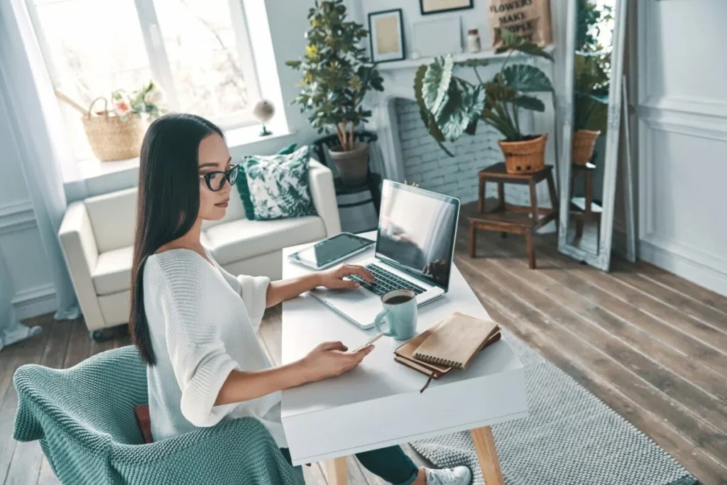 Person working on a laptop at a home office desk in a bright living space with plants and natural light.