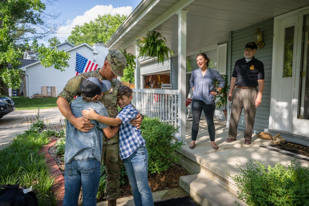 Service member embracing children outside a family home while relatives stand on the front porch nearby.