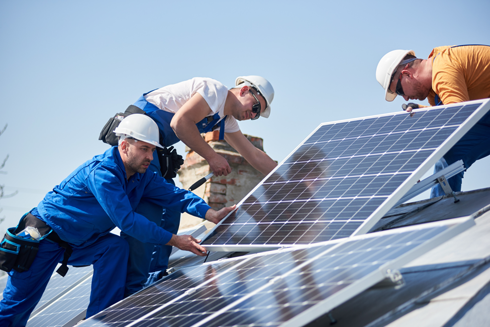 Three workers in safety helmets and uniforms installing solar panels on a sloped rooftop under a clear blue sky.