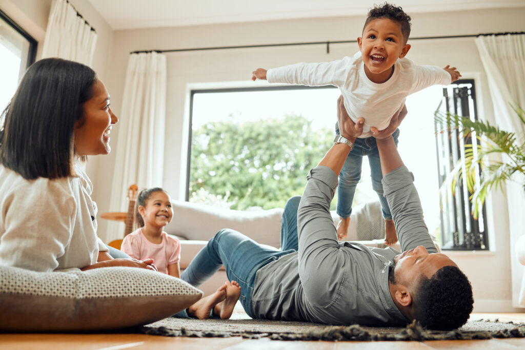 Family, father lifting son and playing in living room with fun and mother with daughter bonding at home together. Happy people with love, care and carefree, airplane and parents relax with kids