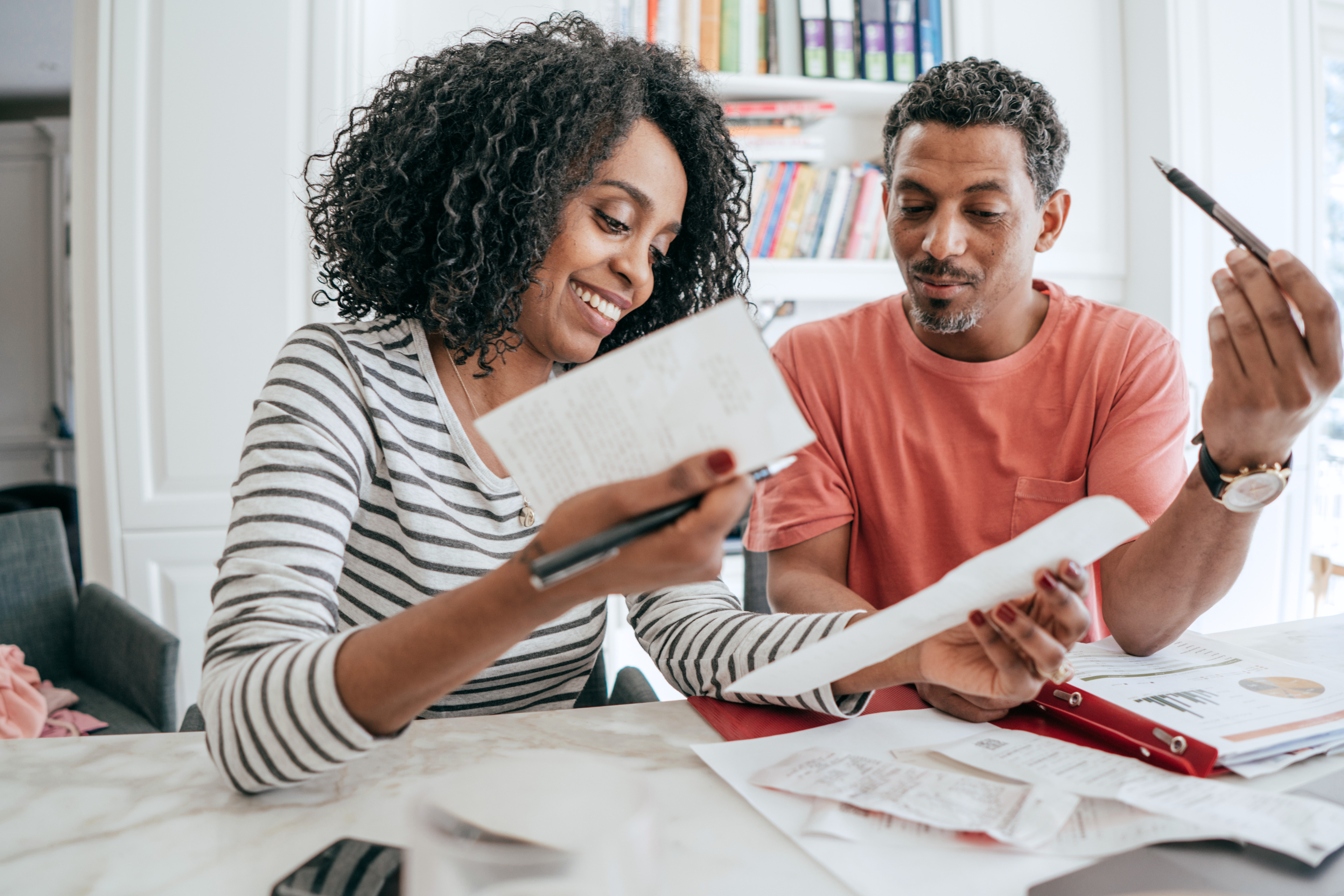 Couple Reviewing Finances Together A smiling couple sits at a table reviewing paperwork and receipts. The woman, holding a pen and notebook, looks happy while discussing the documents. The man, also holding a pen, looks engaged in the conversation. Papers and a binder are spread out on the table in front of them.