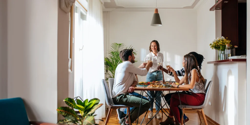 Group gathered around a dining table in a bright home, sharing a meal and raising glasses during a housewarming gathering.
