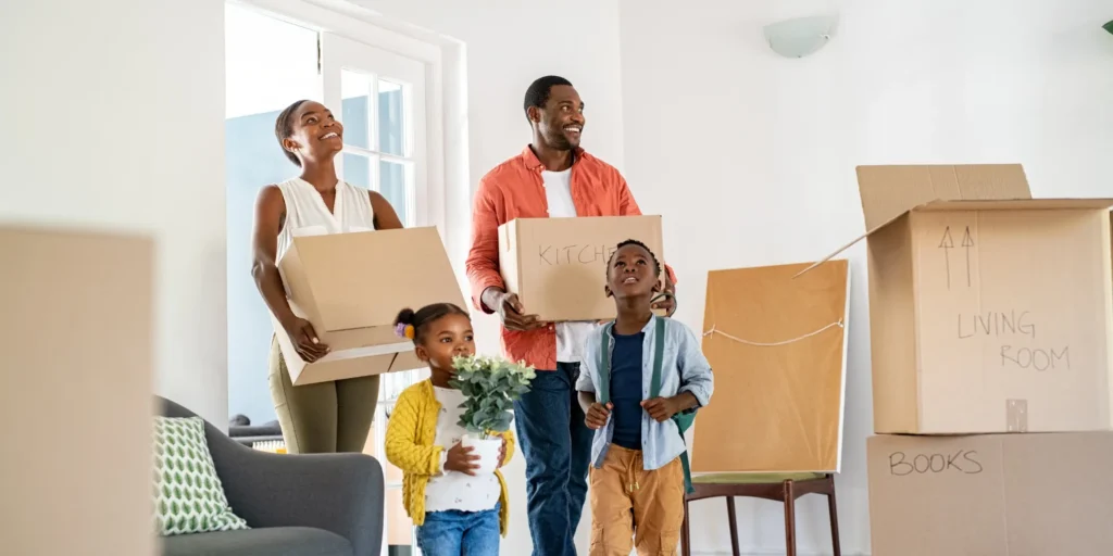 Family carrying moving boxes into a new home, with packed boxes labeled “Kitchen,” “Living Room,” and “Books” visible.