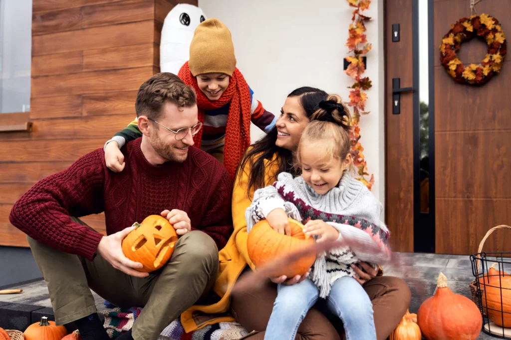 Group of four people carving pumpkins together on a front porch decorated with pumpkins and autumn accents.